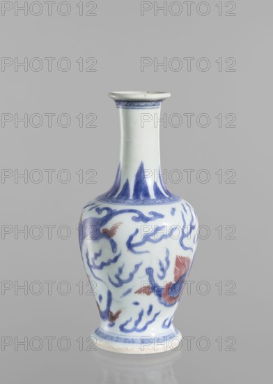 Vase, blue and white porcelain, Chinese art, Qing style, Chinese culture, pearls, dragons iconography, Qing 1644–1911, 19th century, China, vessel, height 19.6 cm, diameter 9.3 cm, A slender porcelain vase standing on a reflective surface against a plain light gray background, with a narrow flared neck and a rounded body tapering to a slightly flared foot. The vase is predominantly white with painted blue decorative motifs that resemble flowing cloud-like forms and stylized shapes around the neck and body, and a band of blue patterning near the rim and the foot. There are also several reddish-orange accents in the form of feathered or petal-like elements integrated into the painted design. Overall the visible colors are white, blue, reddish-orange, and the light gray of the background.