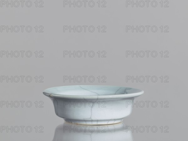 Flowerpot saucer, Chinese ceramics, Qing 1644–1911, 19th century. Grey-blue glaze with cracklework; glazed ceramic vessel, saucer, height 3.5 cm, diameter 11.4 cm., A shallow bowl with a flared rim sits on a reflective surface, showing a smooth, glazed finish with visible crackle lines; the bowl is primarily pale blue with subtle variations of light blue and hints of white, and there are small areas of warm beige near the base and a tiny spot on the rim, all against a uniform gray background.