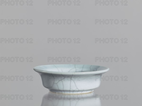 Flowerpot saucer;, Chinese ceramics, Qing dynasty 1644–1911, 19th century; glazed cracklework grey-blue glaze with cracklework; vessel; height 3.5 cm, diameter 11.4 cm., A shallow glazed ceramic bowl with a flared rim sits centered on a reflective surface; the bowl is pale blue with a network of darker crackle lines and subtle variations of light blue and gray across its surface, and a thin band of off-white or cream is visible at the base where it meets the reflection; the background is a uniform light gray.