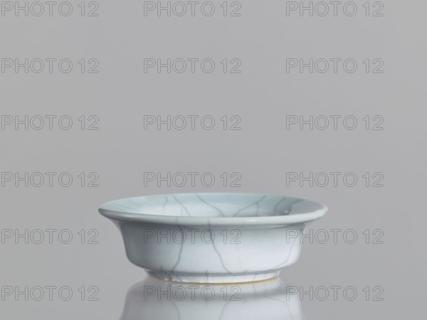 Flowerpot saucer, Chinese ceramics, Qing 1644–1911, 19th century. Grey-blue glaze with cracklework. Porcelain vessel, saucer; height 3.5 cm, diameter 11.4 cm., A shallow ceramic bowl with a flared rim resting on a reflective surface, showing a smooth glazed finish with visible crackle lines; the bowl and its reflection display pale blue and light gray tones with subtle off-white and cream accents, and the background is a uniform light gray.