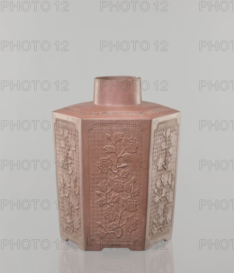 Yixing clay storage vessel; maker, red-brown stoneware with plastic, impressed decoration; decorated with peonies; Qing dynasty 1644–1911, 19th century; Yixing style; Chinese; vessel; height 22.4 cm, width 17.7 cm, depth 15.7 cm, weight 1.15 kg., A hexagonal ceramic vessel with a short cylindrical neck, shown against a pale gray background. The vessel is predominantly reddish-brown with lighter beige accents along the edges of its panels. Each of the visible vertical panels features raised floral reliefs with petals and leaves, rendered in the same reddish-brown and lighter beige tones. The top surface around the neck shows a subtle decorative band in the reddish-brown color. The base has small notches and rests on a reflective surface that mirrors the colors and shapes faintly.