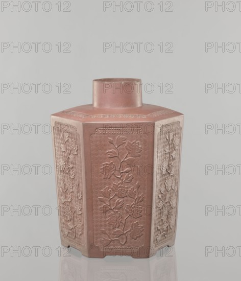 Yixing clay storage vessel, gift; Chinese, Qing dynasty 1644–1911, 19th century. Red-brown stoneware with plastic, impressed decoration depicting peonies. Vessel, height 22.4 cm, width 17.7 cm, depth 15.7 cm, weight 1.15 kg., A hexagonal vase with a short cylindrical neck sits against a plain light gray background, the vase displaying a muted reddish-brown surface with lighter beige highlights on its angled sides; each flat panel features a raised floral relief showing stems, leaves, and blossoms, and the top rim and shoulder have subtle decorative borders, while the vase rests on a reflective surface that produces a faint mirrored outline.