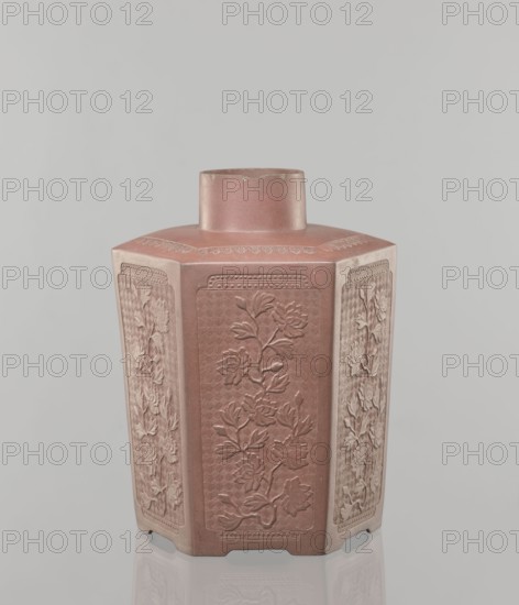 Yixing clay storage vessel, maker, ; Chinese; Qing dynasty 1644–1911; Yixing style; red-brown stoneware with plastic, impressed decoration; subject: peonies; 19th century, China; vessel; height 22.4 cm, width 17.7 cm, depth 15.7 cm, weight 1.15 kg, A hexagonal ceramic vessel stands upright against a pale gray background, showing a muted reddish-brown surface with lighter, dusty highlights. The vessel has a short cylindrical neck rising from a flat top and each vertical face features a recessed panel with raised floral reliefs depicting stems, leaves, and blossoms. The panels vary slightly in tone, with some areas appearing more pinkish and others leaning toward tan or beige, and fine decorative borders surround each panel. The overall finish is matte with subtle variations in texture, and a soft reflection of the base is visible on the surface beneath.