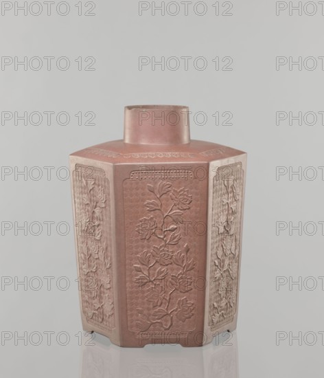 Yixing clay storage vessel, gift; Chinese, Qing dynasty 1644–1911, 19th century. Red-brown stoneware with plastic, impressed decoration depicting peonies. Vessel, height 22.4 cm, width 17.7 cm, depth 15.7 cm, weight 1.15 kg., A hexagonal ceramic vase with a short cylindrical neck, shown against a plain light gray background; the vase has a muted reddish-brown color with lighter beige highlights on some faces, and each vertical panel is decorated with raised floral reliefs depicting stylized blossoms and leaves, with decorative borders at the top and bottom; the surface shows subtle texture and slight variations in color, and the vase stands on a reflective pale surface that creates a faint mirrored reflection.