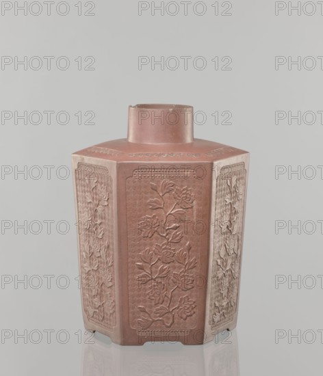 Yixing clay storage vessel, gift, Chinese art, Chinese, Qing 1644–1911, Yixing style, container with peony decoration, red-brown stoneware with plastic, impressed decoration, 19th century, China, vessel; height 22.4 cm, width 17.7 cm, depth 15.7 cm, weight 1.15 kg, A hexagonal ceramic vessel with a short cylindrical neck, shown against a pale gray background; the vessel is primarily a muted reddish-brown with areas of lighter, dusty beige on some faces and edges, and each visible panel features raised floral reliefs depicting stylized blossoms and leaves, with decorative borders framing the panels and a subtle textured pattern behind the floral motifs, and a faint reflection of the base visible on the glossy surface beneath.