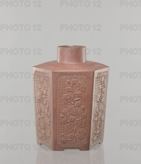 Yixing clay storage vessel, Chinese, Qing dynasty 1644–1911, Yixing style; red-brown stoneware with plastic, impressed decoration depicting peonies; 19th century, China; vessel; height 22.4 cm, width 17.7 cm, depth 15.7 cm, weight 1.15 kg., A hexagonal ceramic vessel with a short cylindrical neck sits centered against a plain light gray background, the vessel showing a reddish-brown color with paler beige highlights on some panels and edges; each visible vertical panel is decorated in low relief with floral motifs and leafy branches, surrounded by patterned borders, and the surface shows subtle textural variations and small scuffs; the base rests on a reflective surface that creates a faint mirrored outline beneath it.