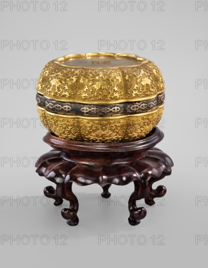 Three-element box on a stand. The box is in the shape of two eight-petal cups connected by a metal rim. Gilded relief decoration of floral tendrils lotuses, chrysanthemums. Wooden round base with a central hole; on five strongly curved legs; side in the shape of a flower rosette broken — three parts. Ceramic. 18th century, China; Qing, Qianlong period 1736–1795. Vessel/container. Box: height 7.8 cm, diameter 13.5 cm, weight 0.73 kg. Stand: height 8.9 cm, diameter 16.0 cm, weight 0.25 kg., function: box., A rounded ornate container with a richly textured gold surface sits atop a dark wooden stand; the container shows intricate raised floral and vine patterns in gold, a darker band encircling its middle with a repeating geometric motif, and a slightly indented lid rim. The wooden stand is polished and dark brown with a carved, scalloped top and curved, bulbous legs. The background is a uniform light gray.