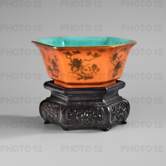 Bowl on a stand. Decoration: peony bushes on an orange background; turquoise bottoms inside and in the mirror. Wooden stand, hexagonal on six legs; carved with openwork. Tongzhi period 1862–1874, China. Porcelain vessel, height 6.8 cm, width 16.6 cm, depth 14.5 cm., An orange hexagonal ceramic bowl with painted floral motifs on its exterior and a turquoise interior sits on a dark carved wooden stand; the bowl has a thin gold rim and the scene shows shades of orange, turquoise, gold, dark brown, black, and gray.