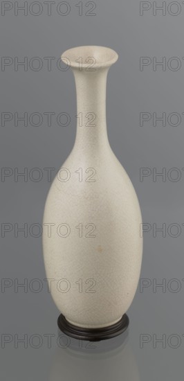 Vase on saucer. Vase made of stoneware or thick porcelain?, ovoid with a long cylindrical neck, flaring at the spout. Light cream white matte glaze covered with fine cracklework. 19th century, Japan, Edo 1603–1868. Ceramics; cracklework decoration; bottles. Vessel. Height 24.1 cm, diameter 8.6 cm, weight 0.65 kg., A tall, narrow-necked ceramic vase with a flared rim stands on a dark round base; the vase surface is a pale off-white or cream with a fine crackle pattern and a few small darker specks and slight discolorations, and the background is a uniform mid-gray.