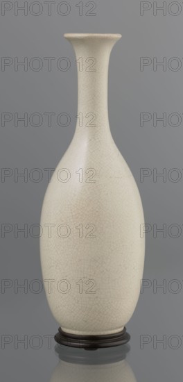 Vase on saucer. Stoneware or thick porcelain?, ovoid with a long cylindrical neck flaring at the spout; light cream white matte glaze covered with fine cracklework. Bottle-type vessel, 19th century, Japan Edo period influence. Japanese ceramics; height 24.1 cm, diameter 8.6 cm, weight 0.65 kg., A tall, slender vase with a narrow neck and flared rim sits on a dark round stand; the vase surface is an off-white or cream color with a visible fine crackle pattern and subtle beige and light brown variations, set against a soft gray background and reflected faintly below.