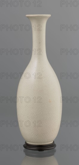 Vase on saucer. Vase made of stoneware or thick porcelain?, ovoid with a long cylindrical neck flaring at the spout; light cream white matte glaze covered with fine cracklework. 19th century, Japan, Edo 1603–1868. Ceramic vessel, cracklework decoration, bottles. Height 24.1 cm, diameter 8.6 cm, weight 0.65 kg., An elongated ceramic vase with a narrow neck flaring to a slightly wider rim, standing on a dark round wooden base; the vase surface is an off-white or cream color with a fine network of hairline crackle patterns in subtle gray and faint brown tones, and there are a few small darker spots and slight warm-beige discolorations on the body; the background is a smooth mid-gray.