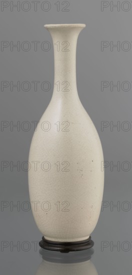 Vase on saucer. Vase made of stoneware or thick porcelain?, ovoid with a long cylindrical neck flaring at the spout; light cream white matte glaze covered with fine cracklework. Bottle-shaped vessel with saucer; ceramics; Japanese, Edo 1603–1868; 19th century. Height 24.1 cm, diameter 8.6 cm, weight 0.65 kg., A tall, narrow-necked ceramic vase with an elongated, rounded body and a flared rim sits on a small dark stand; the vase surface is an off-white or cream color with a fine network of crackled lines and a few small darker spots, and the background is a smooth gray.