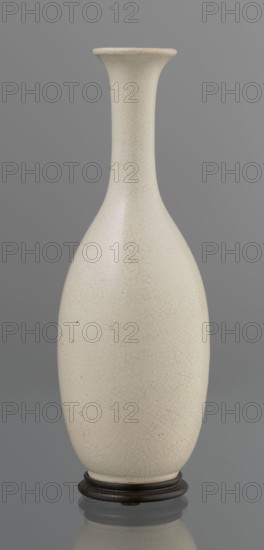 Vase on saucer. Vase made of stoneware or thick porcelain?, ovoid with a long cylindrical neck flaring at the spout; light cream white matte glaze covered with fine cracklework. Bottle-form vessel with saucer; 19th century, Japan, Edo 1603–1868. Ceramics; cracklework decoration. Height 24.1 cm, diameter 8.6 cm, weight 0.65 kg., A tall, narrow-necked vase with a flared rim standing on a round dark wooden base; the vase surface is off-white or cream with a fine crackle pattern and a few small dark specks, and the background is a uniform gray.