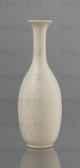 Vase on saucer. Vase made of stoneware or thick porcelain?, ovoid with a long cylindrical neck flaring at the spout. Light cream white matte glaze covered with fine cracklework. 19th century, Japan, Edo 1603–1868. Ceramics; bottles. Vessel. Height 24.1 cm; diameter 8.6 cm; weight 0.65 kg., A pale off-white ceramic vase with a long narrow neck flaring slightly at the rim and a rounded body, showing a fine crackle pattern across its surface and a few small dark specks, set against a smooth mid-gray background with a subtle reflection of the vase at the base; visible colors include off-white, light beige in the crackle lines, dark tiny specks, and gray in the background and reflection.