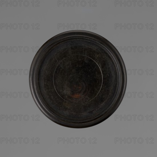 Vase on saucer. Vase made of stoneware or thick porcelain?, ovoid with a long cylindrical neck, flaring at the spout. Light cream white matte glaze covered with fine cracklework. 19th century, Japan, Edo 1603–1868. Ceramics; bottles/vessel. Height 24.1 cm, diameter 8.6 cm, weight 0.65 kg., A circular, dark brown to black object with a raised rim sits centered against a flat gray background; the object's surface shows a worn, slightly scratched texture with faint concentric circles and a small area of subtle reddish-brown discoloration.