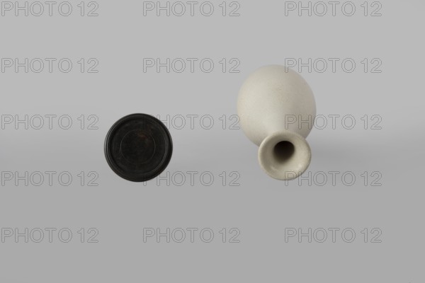 Vase on saucer. Vase made of stoneware or thick porcelain?, ovoid with a long cylindrical neck flaring at the spout; light cream white matte glaze covered with fine cracklework. 19th century, Japan, Edo 1603–1868 period. Ceramics; bottles/vessel. Height 24.1 cm, diameter 8.6 cm, weight 0.65 kg., A light gray background with a dark circular object on the left and a light beige, rounded vase-like object on the right; the dark object appears black with subtle brownish and grayish wear, while the vase shows a textured, off-white to cream surface with darker shadowing inside its hollow opening.