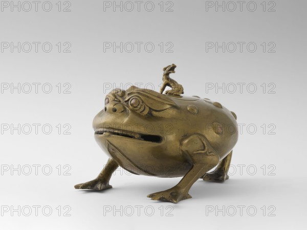 Incense burner with a cover — moon frog toad. Cast in bronze studded with copper. Popular motif in Chinese geomancy — symbol of a three-legged frog toad bringing money wealth. 19th century, Burma Myanmar. Burmese art; ceremonial vessel; frog, wealth/luck and coin/tiger symbolism. Vessel incense burner. Height 20.0 cm; width 32.0 cm; depth 21.0 cm., A metallic sculpture of a frog-like creature in a golden-bronze color sits on a light gray surface against a pale gray background, with a smaller similar creature mounted on its back; the larger creature has a rounded, bulbous body with raised bump details, pronounced eyes, a wide mouth with a narrow opening, short sturdy legs ending in webbed feet, and carved decorative lines on its face and limbs, while the smaller figure is similarly colored and posed with an open mouth.