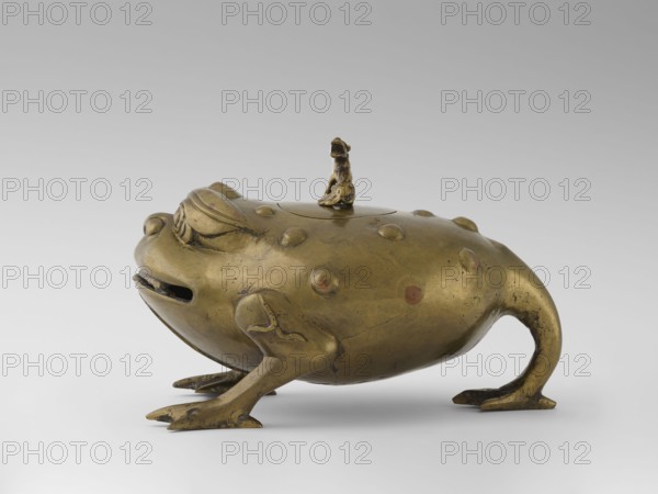 Incense burner with a cover — moon frog toad. Cast in bronze studded with copper. Popular motif in Chinese geomancy: a three-legged frog toad bringing money wealth. 19th century, Burma Myanmar. Burmese art; ceremonial vessel; incense burner. Symbols: frogs, coins, tiger luck/wealth. Height 20.0 cm; width 32.0 cm; depth 21.0 cm., A metallic sculpture of a stylized frog-like creature with a rounded body and smooth surface, shown in a side view. The creature has a pronounced snout with a horizontal opening that resembles a mouth, raised bulging eyes, and several raised rounded bumps on its back. It stands on four limbs with webbed, splayed feet, the rear limbs curved and extending backward. A small sculpted figure sits on a circular lid centered on the creature's back. The surface shows a warm golden-bronze color with darker areas of patina and a few subtle reddish-brown spots. The background is a smooth gradient from light gray at the bottom to a slightly darker gray at the top.