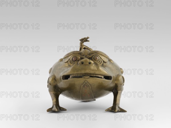 Incense burner with a cover — moon frog toad. Cast in bronze studded with copper. Popular motif in Chinese geomancy — symbol of a three-legged frog toad bringing money wealth. 19th century, Burma Myanmar. Burmese art; ceremonial vessel; incense burner. Motifs: frogs, coins, tiger; symbols of wealth and luck. Height 20.0 cm; width 32.0 cm; depth 21.0 cm., A metallic sculptural object shaped like a squat creature with a rounded body, short legs, and a wide mouth is shown from the front; the surface appears largely brass-colored with areas of darker patina and hints of reddish-brown on the eyes and some worn spots. The creature has stylized facial features including prominent brows, circular eyes set into carved sockets, a small raised nose area with visible nostril openings, and a horizontal mouth opening with a suggestion of teeth. A small ornamental figure sits atop the creature’s head. The legs end in flattened, webbed-looking feet that rest on a pale, neutral background that transiti...