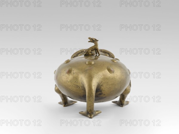 Incense burner with a cover - moon frog toad. Cast in bronze studded with copper. Popular motif in Chinese geomancy — symbol of a three-legged frog toad bringing money wealth. 19th century, Burma Myanmar. Burmese art; ceremonial vessel; incense burner. Subjects/symbols: frogs, coins, tiger; wealth, luck. Height 20.0 cm, width 32.0 cm, depth 21.0 cm., A rounded metallic vessel with a golden-bronze color sits on a flat surface, supported by a group of four short, tapered legs that end in flattened, triangular feet; the vessel’s domed body shows a few raised circular bumps and surface scratches and darker areas of patina, and a small sculpted animal figure in the same golden-bronze color is mounted on top, facing outward.