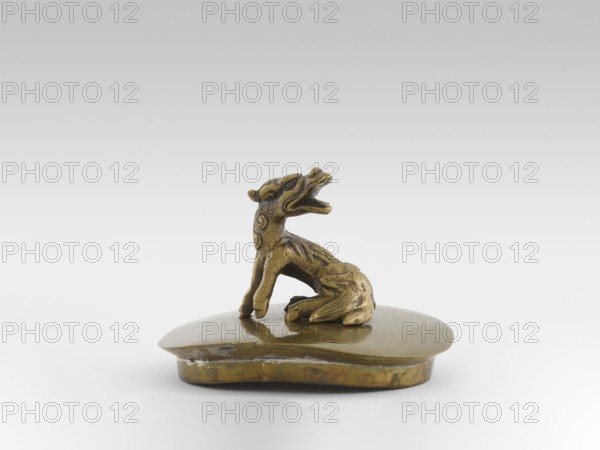 Incense burner with a cover shaped as a moon frog toad. Cast in bronze studded with copper. Popular motif in Chinese geomancy — three-legged frog toad symbolizing the bringing of money/wealth; motifs include frogs, coins and a tiger symbol of luck/wealth. 19th century, Burma Myanmar. Ceremonial vessel, incense burner. Height 20.0 cm, width 32.0 cm, depth 21.0 cm., A small metallic figurine in golden and bronze tones sits on a flat rounded base; the figure resembles an animal with a stylized head held upward and an open mouth, a curved body with carved decorative swirls on its neck, front legs positioned forward, and a tail curled alongside its hindquarters, all showing areas of darker patina and shine against the smoother surface of the base, with the background appearing pale gray and white.