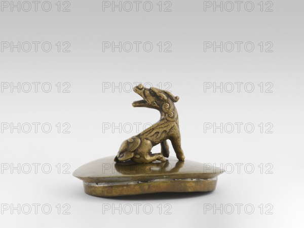 Incense burner with a cover — moon frog toad. Cast in bronze studded with copper. Popular motif in Chinese geomancy — symbol of a three-legged frog toad bringing money wealth. 19th century, Burma Myanmar. Burmese art; ceremonial vessel; incense burner. Symbols: frogs, coins, tiger wealth, luck. Height 20.0 cm; width 32.0 cm; depth 21.0 cm., A small carved object in brass tones sits on a low, rounded base of similar brass color; on top of the base a stylized animal figure with a raised head and open mouth is depicted, showing curved decorative grooves and a textured surface, with the figure's body and limbs forming a compact posture and a tail that curls slightly; the overall color palette includes shades of gold, brass, and muted brown against a pale gray to white gradient background.