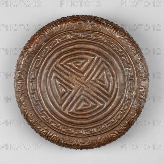 Box, Himalayan, Tibetan, 19th–20th century; container; height 3.8 cm, diameter 12.6 cm., A round, slightly domed metal object with a central raised geometric pattern featuring interlocking triangular and linear shapes, surrounded by concentric raised rings and a decorative border with repeating carved motifs; the surface shows a brownish metallic color with areas of darker brown patina and small flecks of greenish oxidation, set against a pale gray background.