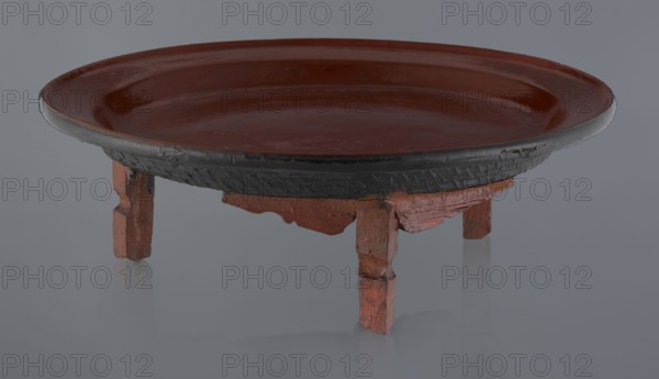 Ritual vessel, Burmese, 19th/20th century. Tray with a fluted surface showing a visible bamboo weaving pattern, made of bamboo covered with black lacquer on the bottom; profiled legs covered with red lacquer. Overall good condition; local abrasions and cracks in the lacquer layers, many local cracks and lacquer losses on the bottom. Height 15.5 cm, diameter 44.5 cm., A shallow round dish with a glossy reddish-brown interior and rim sits on a low, carved stand; the dish has a dark, nearly black outer edge with textured detailing, and the stand beneath is painted in a weathered reddish color with visible wear and rough carved legs.