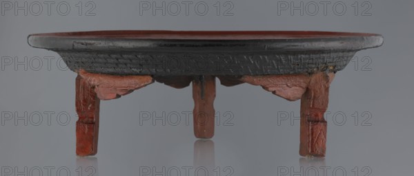 Ritual vessel, Burmese, 19th/20th century. Tray with a fluted surface showing a visible bamboo weaving pattern, made of bamboo covered with black lacquer on the bottom. Profiled legs covered with red lacquer. Used in religious rites. Overall good condition; local abrasions and cracks in the lacquer layers, many local cracks and lacquer losses on the bottom. Height 15.5 cm; diameter 44.5 cm., A low round wooden stand with a wide shallow top and short legs is shown from the front; the top rim and underside are dark black with a textured pattern, while the upper surface and legs are reddish-brown with visible worn and chipped areas; the background is a smooth gray and the stand casts a faint reflection on the surface below; the wood shows uneven surfaces, rough edges, and areas where the black and reddish-brown finishes overlap.