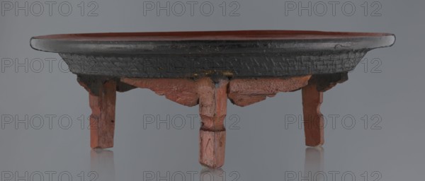 Ritual vessel, gift, Burmese art, Burmese culture, religious rites. Tray with a fluted surface showing a visible bamboo weaving pattern, made of bamboo covered with black lacquer on the bottom. Profiled legs covered with red lacquer. Overall good condition; local abrasions and cracks in the lacquer layers, many local cracks and lacquer losses on the bottom. 19th/20th century, Burma Myanmar. Vessel; height 15.5 cm, diameter 44.5 cm., A low round wooden stand with a shallow rim sits against a neutral background, showing a worn dark top edge and an inner surface with muted reddish-brown tones; the sides beneath the rim display a textured dark finish with areas of wear revealing reddish-brown wood. The stand rests on a group of short carved legs and small supporting brackets with visible tool marks and chipped edges, all in shades of reddish-brown and darkened spots. The background is a uniform gray and the surface beneath the stand reflects the bottom of the legs faintly.