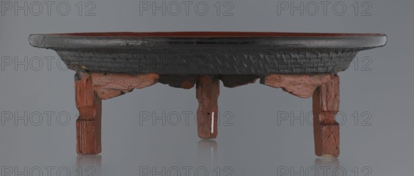 Ritual vessel, Burmese, 19th/20th century. Tray with a fluted surface showing a visible bamboo weaving pattern; made of bamboo covered with black lacquer on the bottom. Profiled legs covered with red lacquer. Overall good condition; local abrasions and cracks in the lacquer layers, many local cracks and lacquer losses on the bottom. Height 15.5 cm; diameter 44.5 cm., A low wooden stand with a rounded top and carved legs is shown against a plain gray background, the top surface appearing dark brown to black with a textured band around its edge, and the inner top surface showing a reddish brown. The legs and supporting brackets are reddish brown with visible wood grain and worn, chipped areas exposing lighter brown tones. The overall colors visible are gray in the background, dark brown or black on the rim, reddish brown on the top interior and legs, and some lighter brown in worn spots.