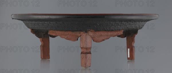 Ritual vessel, Burmese, 19th/20th century. Tray with a fluted surface showing a visible bamboo weaving pattern; made of bamboo covered with black lacquer on the bottom. Profiled legs covered with red lacquer. Overall good condition with local abrasions and cracks in the lacquer layers; many local cracks and lacquer losses on the bottom. Vessel, height 15.5 cm, diameter 44.5 cm., A low wooden stand with a round dark top and a textured black underside rests on a group of reddish-brown carved legs and supports; the top edge shows a thin band of dark brown, the underside surface has a rough patterned texture, and the wooden legs exhibit worn reddish-brown paint with visible chips and exposed wood, all set against a neutral gray background with subtle reflections on a smooth surface below.