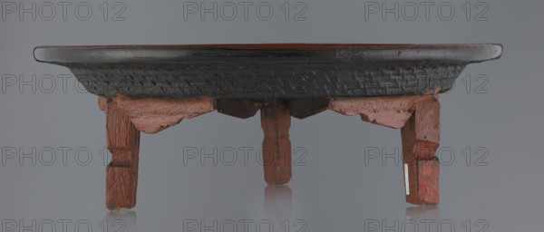Ritual vessel, gift; Burmese art, Burmese culture, religious rites. Tray with a fluted surface showing a bamboo weaving pattern, made of bamboo covered with black lacquer on the bottom. Profiled legs covered with red lacquer. Overall good condition; local abrasions and cracks in the lacquer layers, many local cracks and lacquer losses on the bottom. 19th/20th century, Burma Myanmar. Vessel; height 15.5 cm, diameter 44.5 cm., A low wooden table seen from the front with a round dark tabletop and a carved dark band underneath, supported by a group of short carved legs and decorative aprons in a reddish-brown wood tone; the surface shows areas of worn dark finish and the wood grain and carving details are visible, with the background and floor appearing in muted gray and reflections of the legs faintly visible on the surface beneath.