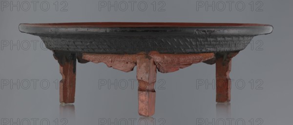 Ritual vessel, gift, Burmese art, Burmese culture, religious rites. Tray with a fluted surface showing a visible bamboo weaving pattern, made of bamboo covered with black lacquer on the bottom. Profiled legs covered with red lacquer. Overall good condition, local abrasions and cracks in the lacquer layers, many local cracks and lacquer losses on the bottom. 19th/20th century, Burma Myanmar. Vessel; height 15.5 cm, diameter 44.5 cm., A low wooden stand with a shallow round top sits on a flat surface, showing a worn rim and textured outer band in dark color and an inner surface in a reddish brown shade, supported by a group of carved legs and brackets in reddish and brown tones with visible chips and rough edges, set against a neutral gray background with subtle reflections on the surface below.