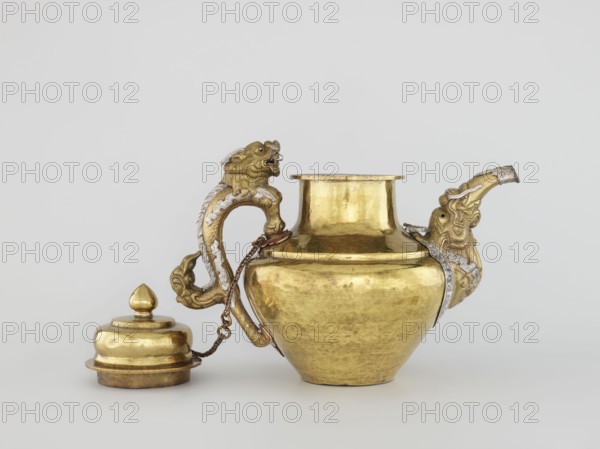 Teapot, Tibetan, 19th century. Jug with a lid on a chain; dragon-shaped handle, spout probably in the shape of a makara. Decorated with dragons, makaras, and lotuses. Repoussé copper sheet, tin-plated inside, bronze-plated outside, patinated; silver-plated in sections. Repoussé spout and handle in low relief with inlaid eyes and copper rivets. Cover topped with a lotus bud. Lhasa. Vessel. Height 25.5 cm; width 31.0 cm; diameter 19.3 cm; weight 0.85 kg., A golden metal vessel with a rounded body and flared neck sits center-right, showing a reflective, slightly worn surface in gold and hints of darker patina; it has an ornate handle shaped like a stylized animal head and curved body with decorative raised detailing in gold and silver tones, and a spout on the right that resembles an animal head with similar gold and silver ornamentation; a lid with a rounded finial in matching gold sits to the left, connected to the vessel by a short metal chain in a darker metallic tone; the background ...