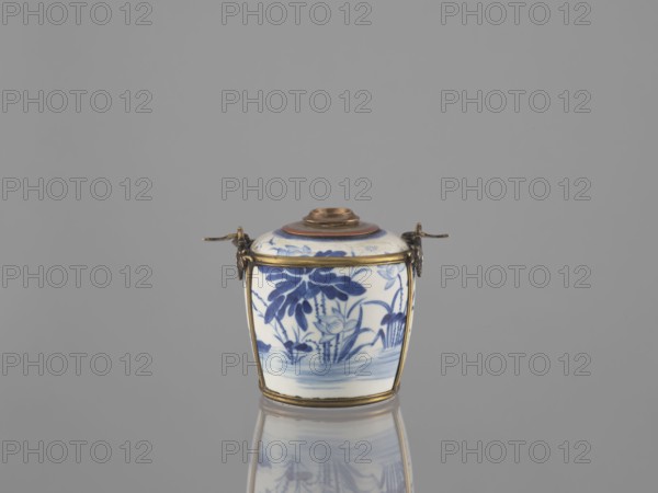 Water pipe, Porcelain belly mounted in a metal frame with floral ornament. Porcelain painted underglaze with cobalt matte glaze. 19th century. Possibly Vietnam or China. Decorated with lotuses and ducks; includes inscriptions. Blue-and-white porcelain smoking pipe. Height 11.0 cm, width 14.6 cm, depth 10.1 cm., A small lidded ceramic container with blue floral and leaf patterns on a white background sits centered on a reflective surface, framed by thin metallic bands and small decorative metal fittings on opposite sides; the lid has a concentric raised center with a brownish tone, and the visible colors include blue, white, gold or brass, brown, and shades of gray in the background and reflection.