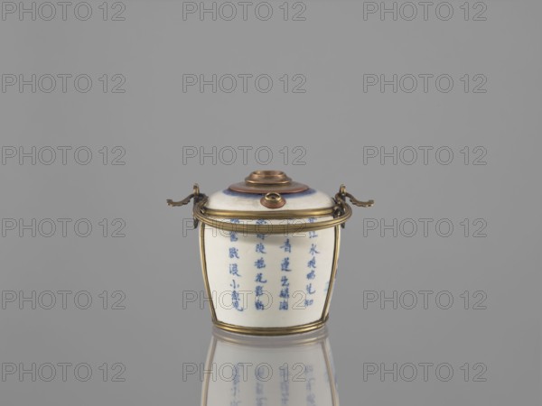 Water pipe, Porcelain belly mounted in a metal frame with floral ornament. Porcelain painted underglaze with cobalt matte glaze. 19th century. Vietnam, China . Blue and white porcelain. Decorations: lotuses, ducks; inscriptions in Chinese. Function: pipe/smoking accessory. Materials: ceramics, metal frame. Dimensions: height 11.0 cm; width 14.6 cm; depth 10.1 cm. Culture/style: Southeast Asian, Vietnamese, Chinese art., A small lidded container sits centered on a reflective surface, showing a mirrored reflection below. The container is predominantly white with blue painted vertical markings on its body, and it is trimmed with metallic gold-colored bands and fittings around the rim and base. The lid has concentric circular details in shades of brown and blue with a small raised central knob in a brass-like color, and two short curved handles on opposite sides with ornate ends in the same metallic gold tone. The background is a smooth uniform gray.