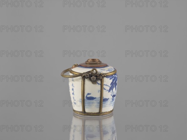 Water pipe, Porcelain belly mounted in a metal frame with floral ornament. Porcelain painted underglaze with cobalt matte glaze. 19th century. Possibly Vietnam or China. Blue and white porcelain. Depicts lotuses and ducks. Smoking accessory; pipe. Inscriptions present. Height 11.0 cm, width 14.6 cm, depth 10.1 cm. Cultures referenced: Southeast Asia, Vietnamese, Chinese., A small cylindrical ceramic container with a rounded lid sits on a reflective surface, showing a porcelain body decorated with blue floral and bird motifs and a few areas of blue brushwork, encased in a brass or gold-toned metal frame with vertical bands and a hinged decorative clasp, and a metal loop handle attached near the top; visible colors include white, blue, gold, brown, and gray in the background and reflection.
