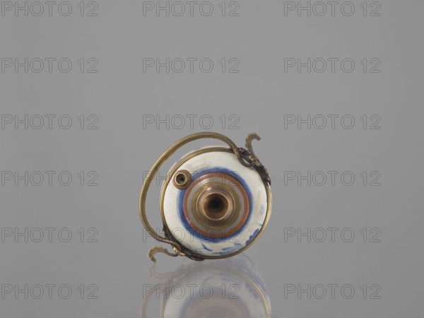 Water pipe, lotuses, ducks, smoking accessories, Southeast Asia culture, ceramics, blue and white porcelain, art of Southeast Asia, Vietnamese culture, inscriptions, Chinese culture, Chinese art. Porcelain belly mounted in a metal frame with floral ornament. Porcelain painted underglaze with cobalt matte glaze. 19th century, Vietnam, China . Pipe. Height 11.0 cm; width 14.6 cm; depth 10.1 cm., A small round object with a glossy white surface and concentric rings in blue and reddish-brown sits centered against a plain gray background, featuring metallic gold-colored fittings and a curved gold handle attached with decorative scrolled mountings, with a small protruding cylindrical opening at the center and a smaller circular fitting nearby, and a soft reflection of the object visible on the surface below; visible colors include gray, white, blue, reddish-brown, and gold.