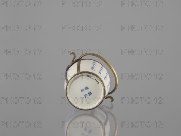 Water pipe, porcelain belly mounted in a metal frame with floral ornament. Porcelain painted underglaze with cobalt matte glaze. 19th century. Subject decoration: lotuses and ducks. Smoking accessory pipe. Materials: ceramics, blue-and-white porcelain, metal mount. Inscriptions: Chinese. Dimensions: height 11.0 cm, width 14.6 cm, depth 10.1 cm. Culture: Southeast Asia Vietnamese?, China., A small white ceramic cup with blue painted designs sits on its side against a smooth gray background, fitted with a gold-colored metal handle and decorative metal bands that curve around the body; a few ornate gold-colored accents are attached where the handle meets the cup, and the cup and metal show a soft reflection on the surface beneath.