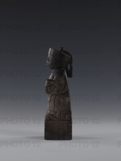 Dignitary, unknown,  craftsman author, Chinese art, Chinese culture, Qing 1644–1911, sages, 19th century, China, figurine, height 19.2 cm, A dark brown carved figure with a rounded head and a cylindrical base stands in profile against a gradient gray background; the figure wears a tall headpiece and a long robe with carved folds, has folded arms across the chest, a pronounced nose and chin, and a textured surface showing lighter brown wear and subtle greenish-gray patina.