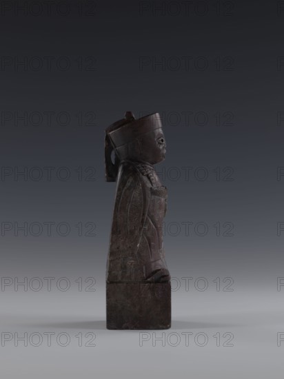 Dignitary, Chinese art, Chinese culture, Qing 1644–1911, sage, 19th century, China, figurine, height 19.2 cm, A dark brown carved figure standing on a rectangular base, shown in profile with a rounded head wearing a tall hat and a strap or ribbon hanging down the back; the figure has a prominent nose and carved facial features, a long draped garment with incised decorative patterns and folded sleeves, and hands positioned near the chest; the base and sculpture surface show lighter brown and grayish wear, and the background is a smooth gradient from dark gray at the top to lighter gray toward the bottom.