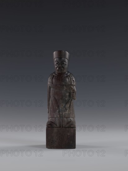 Dignitary, Chinese art, Chinese culture, Qing 1644–1911, 19th century, China, figurine, height 19.2 cm, A dark brown carved wooden figure stands on a square base against a gradient gray background, showing a stylized human form with a cylindrical hat, a rounded face with carved eyes and mouth, a draped garment with incised decorative patterns, and a slightly raised arm detail; colors visible include dark brown on the figure and base, and shades of gray in the background.