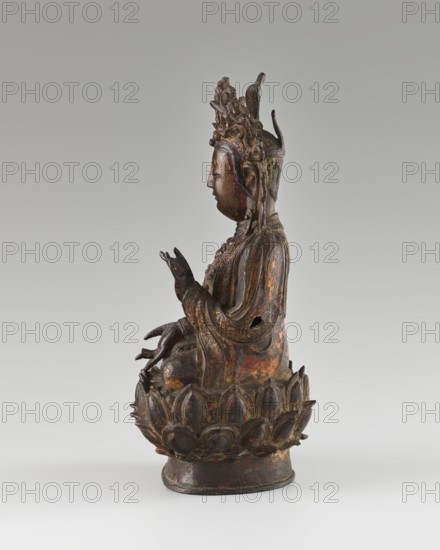 Guanyin, unknown creator; 18th century Qing dynasty 1644–1911, China; Chinese Buddhist sculpture; lotus throne; bodhisattva mercy; figurine; height 24 cm., A three-dimensional sculpture of a seated figure on a lotus-shaped base rendered in dark brown and aged bronze tones with areas of worn reddish and gold highlights; the figure wears an ornate crown with intricate carved details and has long flowing robes with textured folds and layered ornamentation, hands posed in delicate gestures, a serene facial profile with closed eyes, and a small hole visible in the side of the torso; the lotus base shows petal motifs with similar dark brown, bronze, and touches of reddish coloration, all set against a smooth, light gray background.
