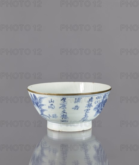 Bowl, Deep round bowl on a high, ring-shaped foot. Decorated with underglaze cobalt painting: exterior shows a stylized composition with elaborate architecture, a human figure, mountains, and an inscription in kanji characters in four columns. Interior undecorated, with a symbol carved into the glaze at the bottom. At the base, a signature reading Chenghua nian zhi—an imitation of a Chinese Ming Dynasty Chenghua reign mark. Glaze has a bluish tint from cobalt; rim secured with a narrow brass ring. Ceramics; figurative scenes; bowls on a foot. Possibly 2nd half of the 17th century, Japan. Height 8.3 cm; diameter 17.7 cm., A small ceramic bowl with a white interior and exterior decorated in blue-painted designs, sitting on a reflective surface that mirrors the bowl's underside and blue patterns; the bowl has a thin metallic rim with a gold tone and a short foot with a slightly discolored base; visible colors include white, blue, gold, and subtle gray tones from the reflection and backgro...