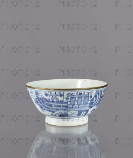 Bowl, Deep round bowl on a high, ring-shaped foot. Decorated with underglaze cobalt painting: outside shows a stylized composition of elaborate architecture, a human figure, and mountains, plus an inscription in kanji in four columns. Interior undecorated, with a symbol carved into the glaze at the bottom. Base bears a signature: Chenghua nian zhi—an imitation of a Chinese Ming Dynasty Chenghua mark. Bluish-tinted glaze from cobalt; rim secured with a narrow brass ring. Edo period 1603–1868; possibly 2nd half of 17th century. Japanese; ceramics, figurative scenes. Vessel height 8.3 cm, diameter 17.7 cm., A small ceramic bowl sits on a reflective surface, showing a mirrored image below. The bowl is primarily white with blue painted decorative patterns around its exterior and a thin gold-colored rim at the top. The interior appears white, and the exterior blue decoration includes a mix of curved and linear motifs. The background is a uniform gray.
