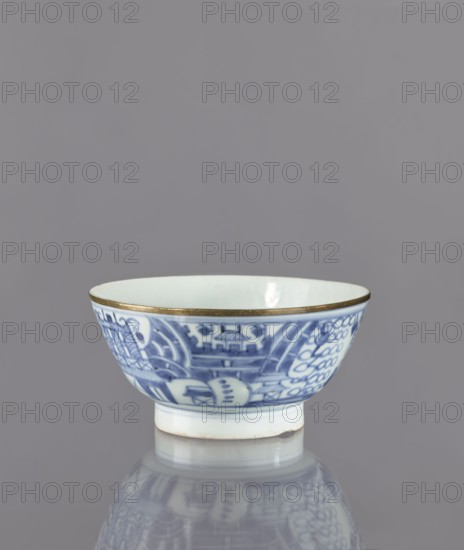 Bowl, Deep round bowl on a high, ring-shaped foot. Decorated with underglaze cobalt painting: exterior shows a stylized composition of elaborate architecture, a human figure, and mountains, with an inscription in kanji in four columns. Interior undecorated, with a symbol carved into the glaze at the bottom. Base bears a signature Chenghua nian zhi — an imitation of a Chinese Ming Dynasty Chenghua reign mark. Glaze has a bluish tint from cobalt; rim secured with a narrow brass ring. Edo period 1603–1868; possibly 2nd half of the 17th century. Japan. Ceramic bowl on a foot; height 8.3 cm, diameter 17.7 cm., A ceramic bowl with blue decorative patterns on a white background and a thin metallic rim sits on a reflective surface, showing a softened mirror image below; the bowl's interior is plain white, and the exterior features intricate blue designs; the visible colors include blue, white, metallic gold, and gray from the background and reflection.