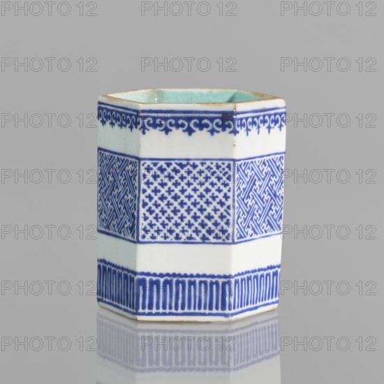 Hexagonal bowl holder in the shape of a prism, incense container. Porcelain body with white glaze outside; inside and foot glazed in turquoise; overglaze decoration with cobalt enamel; bronze foot and rim edge. Decorated with three stripes of borders with geometric ornament meander. Signed. Colored porcelain, geometric ornaments; Qianlong style, Qing dynasty 1644–1911, Qianlong 1736–1795. 19th century . China. Height 7.5 cm, width 5.8 cm. Makership unknown., A hexagonal porcelain vessel standing upright on a reflective surface, predominantly white with blue decorative bands and patterns; the top interior shows a pale green glaze rimmed with a thin brown edge, and the body is divided into horizontal sections featuring a dark blue scrolling border near the top, a band of alternating geometric patterned panels in blue, a plain white band, and a lower blue border of vertical elongated motifs; the background is a neutral gray and the vessel casts a soft reflection beneath it.