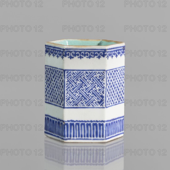Bowl holder, Hexagonal prism-shaped container for incense or bowls, decorated with three stripes of geometric borders and meander ornament. Porcelain body with white exterior glaze, overglaze decoration with cobalt enamel; interior and foot glazed in turquoise; bronze foot and rim edge. Signed. Chinese, Qianlong style Qing dynasty, 1644–1911, 19th century . Height 7.5 cm, width 5.8 cm., A hexagonal ceramic vessel sits on a reflective surface against a gray background, showing white and blue exterior glazing with a pale turquoise interior rim; the sides feature decorative bands including a top scrolling motif in blue, a central section with geometric and lattice-like patterns in blue on white, and a lower band of vertical blue strokes above the base, with subtle signs of wear along the rim and a soft reflection beneath.