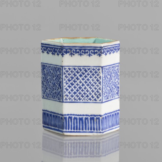 Bowl holder, hexagonal prism-shaped container used as an incense vessel. Porcelain body with white glaze outside, overglaze decoration with cobalt enamel forming three stripes of borders with geometric meander ornament; inside and foot glazed in turquoise. Bronze foot and rim edge. Signed. Chinese; Qianlong style, Qing dynasty 1644–1911, 19th century . Height 7.5 cm, width 5.8 cm., A hexagonal ceramic vessel with a glossy white base and blue decorative patterns sits on a reflective surface against a light gray background; the vessel shows a pale turquoise interior rim, a thin brown edge along the top, bands of intricate blue geometric and scroll-like motifs around its exterior, and a blue patterned border near the base, with reflections of the blue and white visible on the surface below.